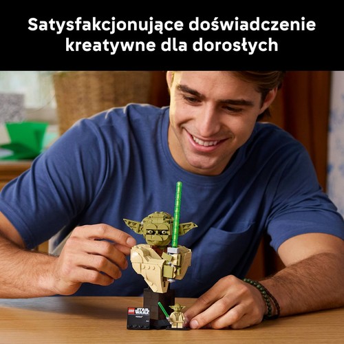 Dorosły mężczyzna z zadowoleniem składa klocki LEGO, tworząc popiersie Yody z uniwersum Star Wars. Model przedstawia Yodę z zielonym mieczem świetlnym, stojącego na czarnej podstawce z plakietką. Obok znajduje się miniaturowa figurka Yody.