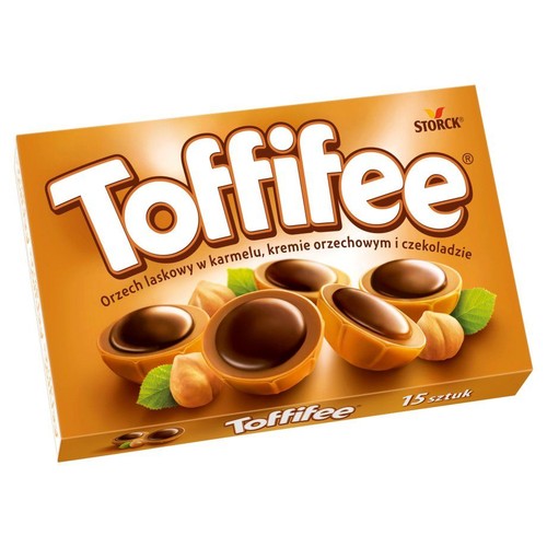 Produkt Toffifee w brązowym pudełku z białym logo. Opakowanie prezentuje pralinki z orzechem laskowym zatopionym w karmelu, otoczone kremem orzechowym i czekoladą. Widoczne są również całe orzechy laskowe i zielone listki. Ilość: 15 sztuk.