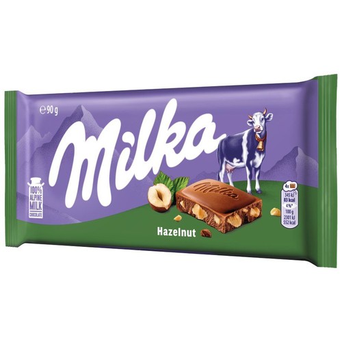 Milka czekolada mleczna z orzechami laskowymi w opakowaniu 90g. Fioletowe tło z górami, zielony dół. Widoczna krowa Milka, kawałek czekolady z orzechami i całe orzechy laskowe.