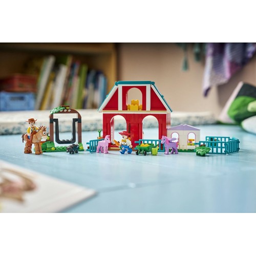 Kolorowy zestaw LEGO Duplo z motywem farmy, rozłożony na niebieskiej podłodze. Zawiera czerwoną stodołę, dwie figurki, w tym kowboja na koniu, dwa różowe konie, czarną świnkę i zielone płotki.