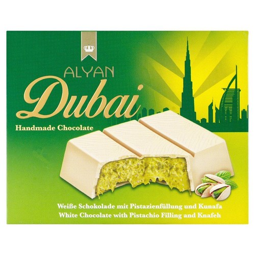 Produkt ALYAN Dubai: ręcznie robiona biała czekolada z nadzieniem pistacjowym i Kunafa. Na zielonym opakowaniu widnieje sylwetka Dubaju. Trzy kawałki czekolady, jeden przełamany, ukazują bogate zielone wnętrze i pistacje.