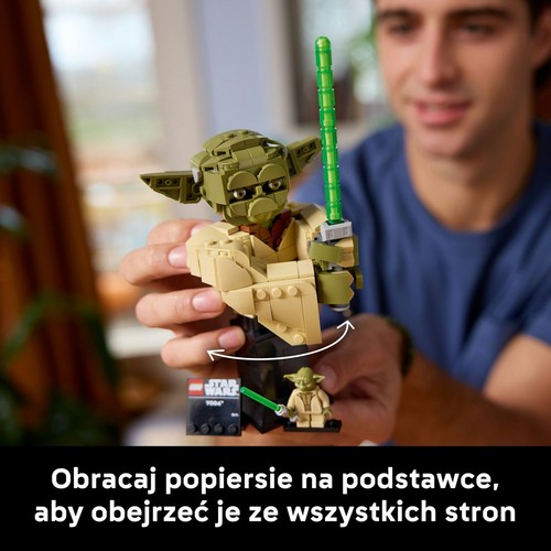 Mężczyzna trzyma w dłoniach dużą, zbudowaną z klocków LEGO figurkę Yody z Gwiezdnych Wojen. Yoda ma zielony miecz świetlny i beżową szatę. Figurka stoi na czarnej, obrotowej podstawce, co pozwala na oglądanie jej z każdej strony.
