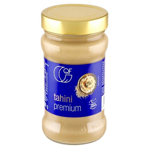Tahini premium QF w przezroczystym szklanym słoiku ze złotą zakrętką. Na niebieskiej etykiecie znajduje się białe logo QF, nazwa produktu "tahini premium" oraz mała grafika dania z sezamem.