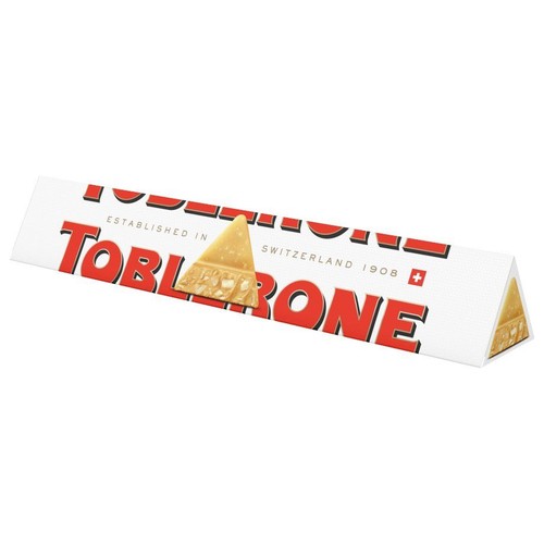 Białe, trójkątne opakowanie czekolady Toblerone. Na froncie duży, czerwony napis "TOBLERONE" oraz mniejszy tekst "Założono w Szwajcarii 1908". Centralnie umieszczona grafika trójkątnego kawałka czekolady z widocznymi kawałkami nugatu.