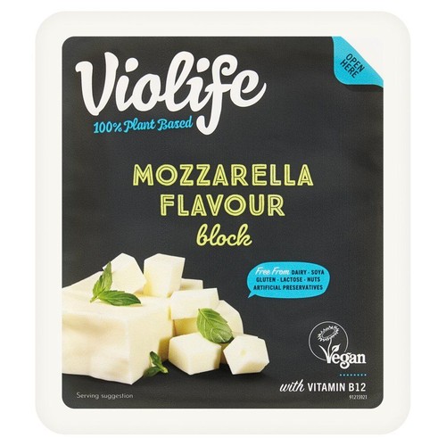 Opakowanie Violife 100% roślinnego bloku o smaku mozzarelli. Na czarnym tle widoczne są białe kostki produktu z listkami bazylii. Produkt jest wegański, bez nabiału, soi, glutenu, laktozy, orzechów i sztucznych konserwantów. Zawiera witaminę B12.