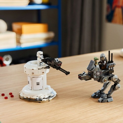 Klocki LEGO Star Wars ułożone na jasnym, drewnianym blacie. Biała wieżyczka z czarnym działkiem i figurką szturmowca stoi po lewej. Po prawej znajduje się szary pojazd kroczący z dwoma figurkami: Mandalorianinem za sterami i małym Grogu z tyłu.