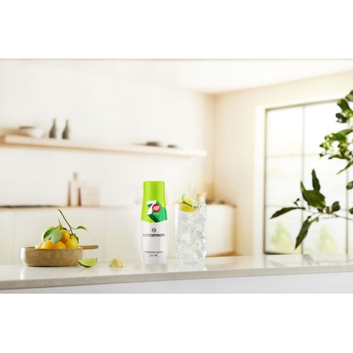 Butelka koncentratu napoju SodaStream 7UP ZERO o pojemności 440 ml, z białą podstawą i zieloną etykietą, stoi na jasnym blacie kuchennym. Obok niej znajduje się wysoka szklanka wypełniona lodem i plasterkami limonki, a także drewniana miska z cytrynami.