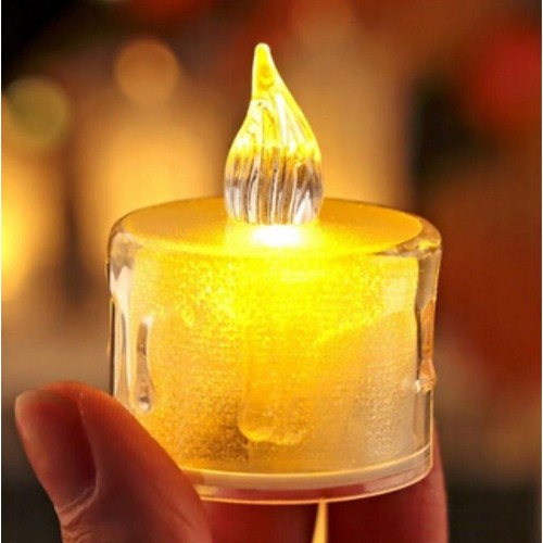 LED-owa świeczka typu tealight z przezroczystą obudową i imitacją kapiącego wosku. Emituje ciepłe, żółte światło, a jej plastikowy płomień realistycznie migocze. Jest trzymana w palcach na rozmytym, ciepłym tle.
