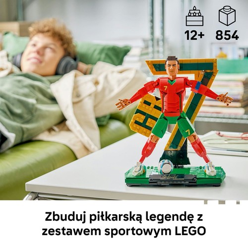 Zestaw LEGO przedstawiający figurkę piłkarza w czerwono-zielonym stroju z numerem 7, stojącą na zielonej podstawce z piłką. Za figurką znajduje się duża, żółta cyfra 7. W prawym górnym rogu ikony: wiek 12+ i 854 elementy.