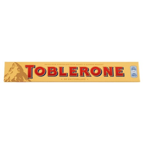 Podłużne, żółte opakowanie czekolady Toblerone. Na opakowaniu widnieje duży, czerwony napis "TOBLERONE" oraz grafika góry. Opis informuje, że to szwajcarska czekolada mleczna z miodem i nugatem migdałowym, pochodząca ze Szwajcarii.