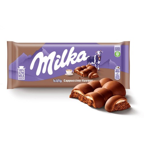 Produkt Milka Bubbly Cappuccino Flavour. Fioletowe opakowanie z napisem Milka, krową i informacją o kakao ze zrównoważonych źródeł. Obok opakowania leżą rozłamane kawałki czekolady z bąbelkowym, jasnym nadzieniem.