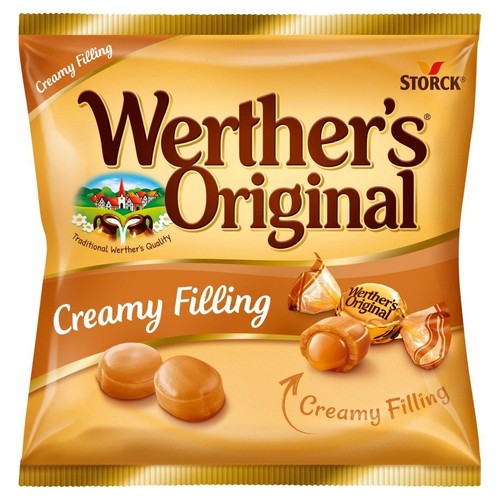 Złota torebka cukierków Werther's Original Creamy Filling. Na opakowaniu widnieje nazwa produktu, logo Storck oraz grafika wsi. Przedstawiono trzy cukierki, w tym dwa rozpakowane z widocznym kremowym nadzieniem.