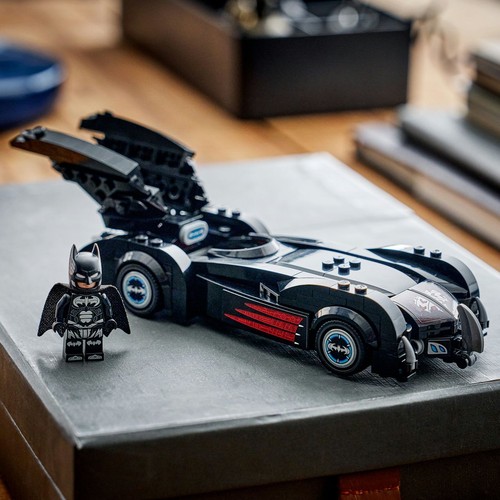 Zestaw LEGO przedstawiający czarny Batmobil z detalami w kolorze czerwonym i niebieskim na kołach, oraz figurkę Batmana w czarnym stroju. Obiekty są umieszczone na szarym podłożu, z widocznym drewnianym blatem w tle.