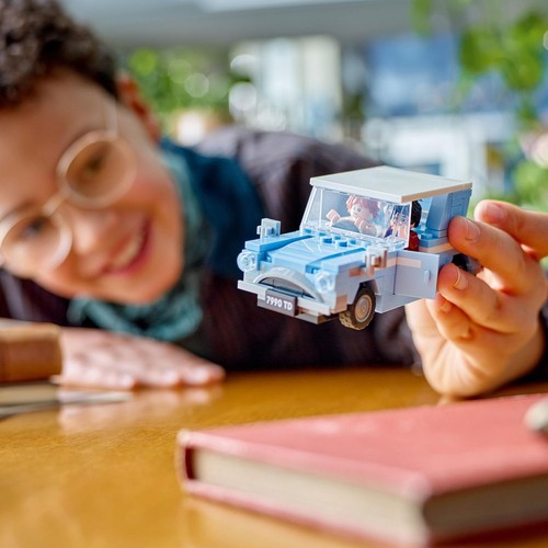 Zbliżenie na dłonie trzymające niebieski samochodzik z klocków LEGO. W samochodzie siedzą dwie małe figurki, a jedne drzwi są otwarte. W tle, poza ostrością, widać uśmiechniętą twarz osoby w okularach oraz fragment czerwonej książki.