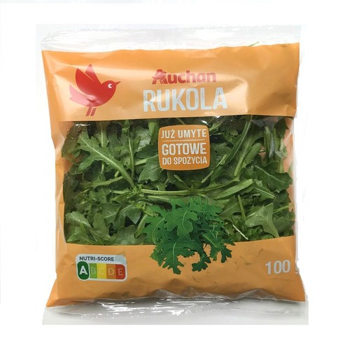 Świeża rukola marki Auchan w przezroczystym woreczku o wadze 100 gramów. Na pomarańczowej etykiecie informacja, że produkt jest już umyty i gotowy do spożycia. Widoczne zielone liście.