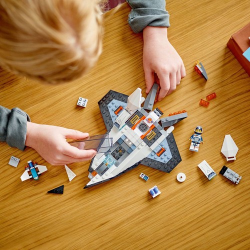 Dziecko z blond włosami bawi się klockami LEGO na jasnym, drewnianym stole. Składa duży, biało-szary statek kosmiczny z pomarańczowymi akcentami. Wokół leżą drobne klocki, mały samolot i figurka astronauty.