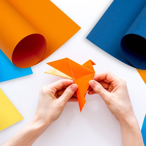 Dwie dłonie precyzyjnie składają arkusz pomarańczowego papieru, tworząc figurkę origami przedstawiającą ptaka. Na białym tle widoczne są również zwinięte rolki pomarańczowego i niebieskiego papieru oraz płaskie arkusze w różnych kolorach.