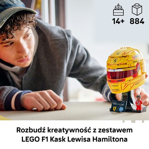 Młody mężczyzna w czapce i bluzie z kapturem patrzy na szczegółowy model LEGO F1 Kask Lewisa Hamiltona. Żółty kask z logo sponsorów stoi na czarnej podstawce z tabliczką i małą figurką. Zestaw zawiera 884 elementy, dla wieku 14+.