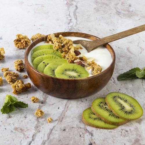 Drewniana miska z jogurtem, plastrami kiwi i granolą. Obok leży drewniana łyżka, kilka kawałków kiwi i listki mięty.
