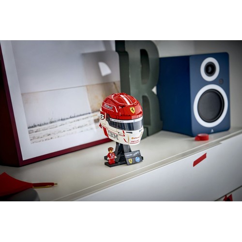Szczegółowy model LEGO kasku wyścigowego Ferrari w barwach czerwono-białych, z numerem 6 i licznymi logo sponsorów, takimi jak IBM, Shell i HP. Kask stoi na czarnej podstawce, obok niego znajduje się mała figurka kierowcy w czerwonym kombinezonie.