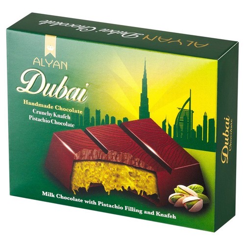 Zielone pudełko czekolady Alyan Dubai z grafiką panoramy Dubaju na żółto-zielonym tle. Na opakowaniu widoczna jest ilustracja przekroju czekolady mlecznej z chrupiącym nadzieniem pistacjowym i knafeh, obok leżą pistacje.