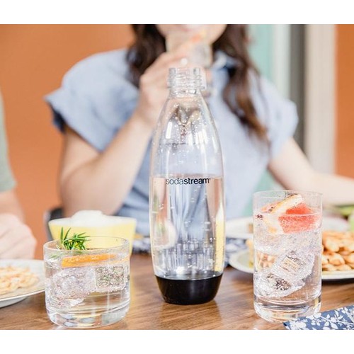 Przezroczysta butelka SodaStream z czarną podstawą, wypełniona wodą gazowaną, stoi na drewnianym stole. Obok niej dwie szklanki z lodem i bąbelkami. Jedna zawiera plaster pomarańczy i gałązkę rozmarynu, druga plaster grejpfruta.