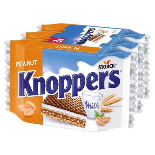 Trzy opakowania wafli Knoppers Peanut ułożone w stos. Na etykiecie widnieje nazwa produktu, grafika przekroju wafla z warstwami, szklanka mleka, orzeszki ziemne oraz kłosy zboża. Opakowanie jest biało-pomarańczowe z niebieskimi elementami.