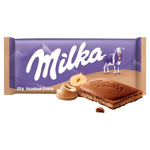 Czekolada Milka 85g z kremem orzechowym w charakterystycznym fioletowym opakowaniu z grafiką krowy. Przed opakowaniem widoczny jest kawałek czekolady z nadzieniem, orzech laskowy oraz porcja kremu.