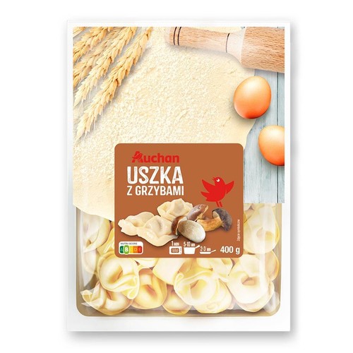 Produkt Auchan Uszka z grzybami 400 g z częściowo przezroczystym opakowaniem. Na brązowej etykiecie widnieje nazwa produktu, logo Auchan i grafika przedstawiająca uszka oraz borowiki i boczniaki. W tle etykiety widoczne są zboże, ciasto i jajka.