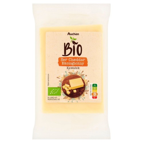 Kawałek sera cheddar ekologicznego marki Auchan BIO w przezroczystym opakowaniu. Widoczny żółty ser. Posiada certyfikat rolnictwa UE.