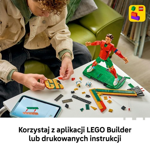 Młoda osoba w beżowej kurtce i ciemnych spodniach składa zestaw LEGO na białym stole. Buduje figurkę piłkarza Cristiano Ronaldo w czerwono-zielonym stroju, stojącą na zielonej podstawie. Obok leży tablet z otwartą aplikacją instrukcji.