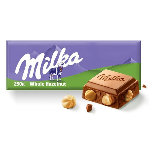 Fioletowe opakowanie czekolady Milka 250g z logo i grafiką krowy. Na opakowaniu napis "Whole Hazelnut". Obok leży odłamany kawałek czekolady mlecznej z widocznymi całymi orzechami laskowymi oraz jeden orzech obok.