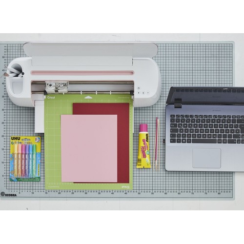 Kompletny zestaw do tworzenia projektów DIY. Zawiera biały ploter tnący Cricut, zieloną matę, dwa arkusze papieru (różowy i czerwony), opakowanie 6 kolorowych klejów brokatowych UHU, żółtą tubkę kleju uniwersalnego UHU, narzędzia i otwarty laptop.