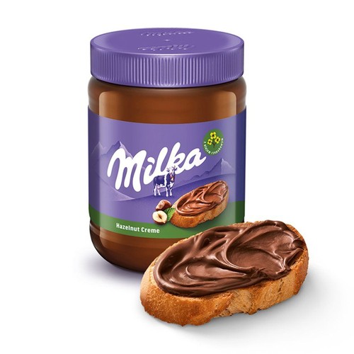 Słoik kremu orzechowego Milka z fioletową etykietą i nakrętką. Na etykiecie logo Milka, krowa, napis "Hazelnut Creme" oraz symbol Cocoa Life. Przed słoikiem kromka pieczywa z apetycznie rozsmarowanym brązowym kremem.