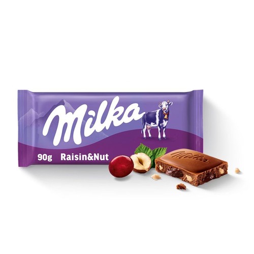 Produkt: czekolada Milka Rodzynka i Orzech 90g. Fioletowe opakowanie z krową. Obok kawałek czekolady z orzechami i rodzynkami, świeże orzechy laskowe, liście i czerwony owoc.