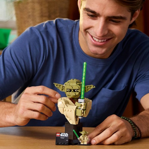 Mężczyzna z zadowoleniem buduje zestaw LEGO Star Wars Yody. Na drewnianym stole znajduje się popiersie Yody z klocków, z zielonym mieczem świetlnym, oraz mniejsza figurka Yody na czarnej podstawce z napisem.