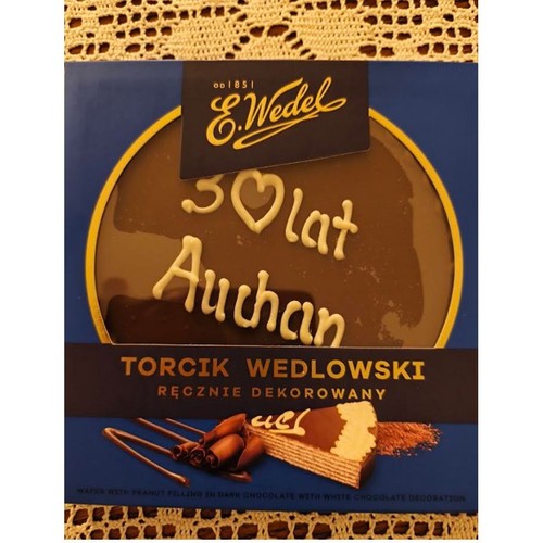 Produkt E. Wedel: Torcik Wedlowski Ręcznie Dekorowany. Na opakowaniu widoczny jest okrągły czekoladowy torcik z białym napisem "30 lat Auchan". Poniżej grafika waflowego ciastka z nadzieniem orzechowym w ciemnej czekoladzie z białą dekoracją.