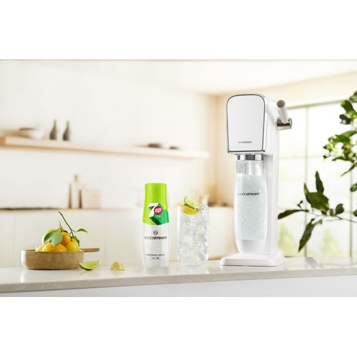 Biały saturator SodaStream z butelką wody gazowanej stoi na jasnym blacie kuchennym. Obok zielono-biała butelka koncentratu napoju 7UP Zero o pojemności 440 ml oraz wysoka szklanka z lodem, napojem i plasterkami limonki. W tle cytryny w misce.