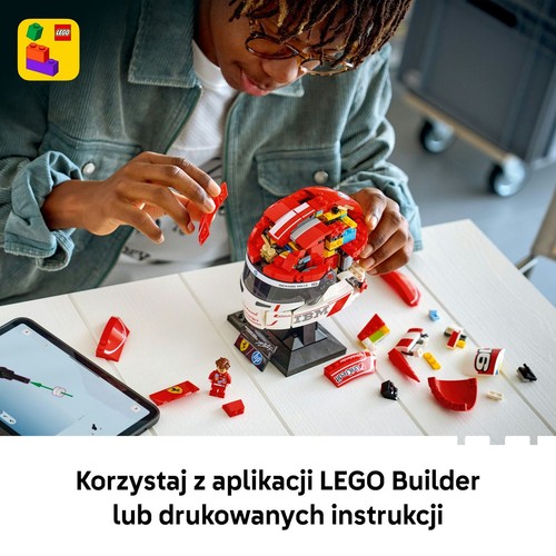 Młody mężczyzna w okularach i dżinsowej kurtce składa model kasku wyścigowego LEGO Ferrari z czerwonych i białych klocków. Kask stoi na czarnej podstawce. Obok leży tablet z cyfrowymi instrukcjami i rozsypane klocki.