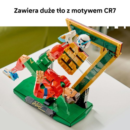 Szczegółowy model LEGO przedstawiający piłkarza w czerwono-zielonym stroju, wykonującego strzał na bramkę. Piłka jest w powietrzu, a figurka stoi na zielonej podstawie. Z przodu znajduje się czarna tabliczka z białym podpisem.