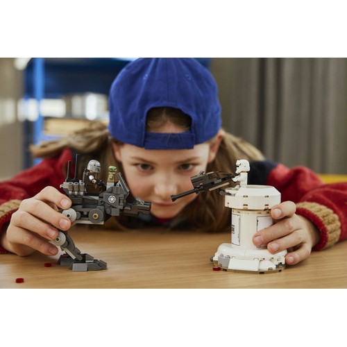 Młoda dziewczynka z bliska, w niebieskiej czapce, intensywnie patrzy na swoje konstrukcje LEGO Star Wars. W rękach trzyma szary AT-ST z dwoma minifigurkami, a obok stoi biała wieżyczka obronna z figurką szturmowca.