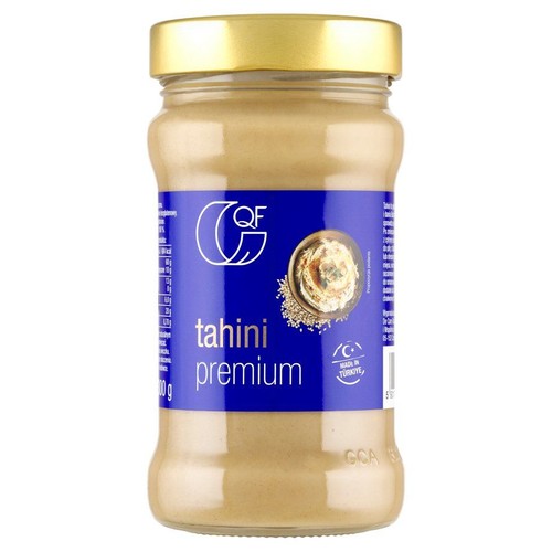 Produkt tahini premium QF w przezroczystym szklanym słoiku o pojemności 400g. Słoik ma złotą zakrętkę i niebieską etykietę z białym napisem "tahini premium". Na etykiecie widoczna jest również grafika potrawy z pastą sezamową.