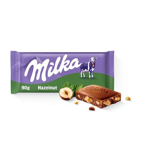 Produkt Milka Hazelnut 90g. Fioletowe opakowanie z logo Milka, wizerunkiem krowy i alpejskim krajobrazem. Przed opakowaniem leży odłamany kawałek czekolady mlecznej z zatopionymi orzechami laskowymi, obok całe orzechy i listki.