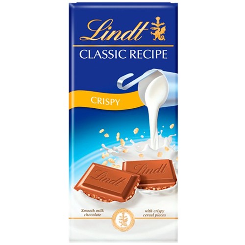 Opakowanie czekolady mlecznej Lindt Classic Recipe Crispy. Granatowe tło z białym logo Lindt. Grafika przedstawia dzbanek wylewający mleko na dwa kawałki gładkiej mlecznej czekolady z chrupiącymi kawałkami zbóż.
