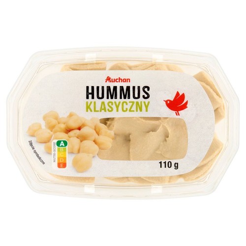 Przezroczyste plastikowe opakowanie hummusu klasycznego marki Auchan, 110 g. Na białej etykiecie widnieje napis "Auchan Hummus Klasyczny" oraz czerwone logo ptaka. W pojemniku widoczny jest kremowy hummus.